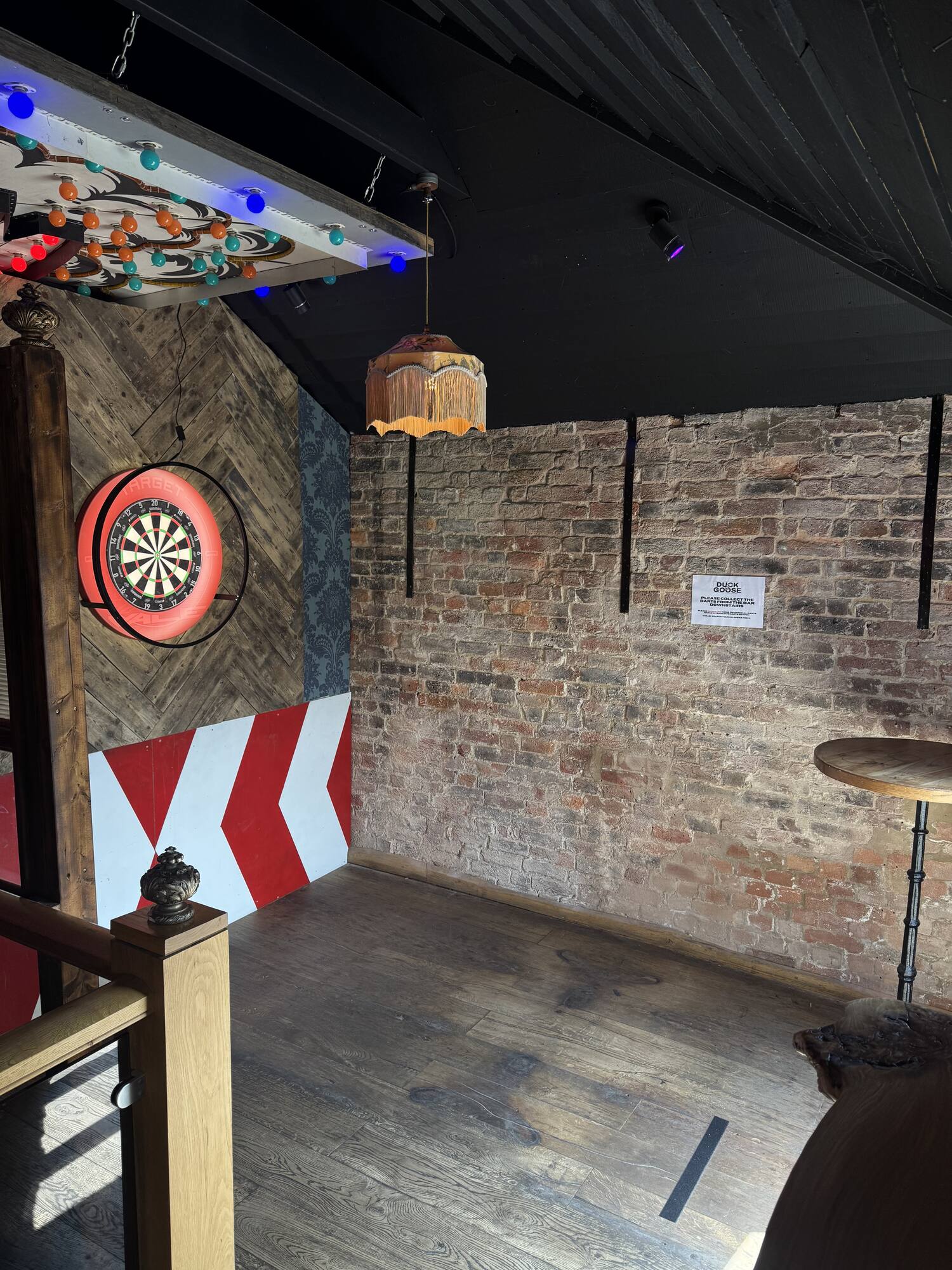 A traditional dartboard and high table on exposed brick, lit by festoon lights