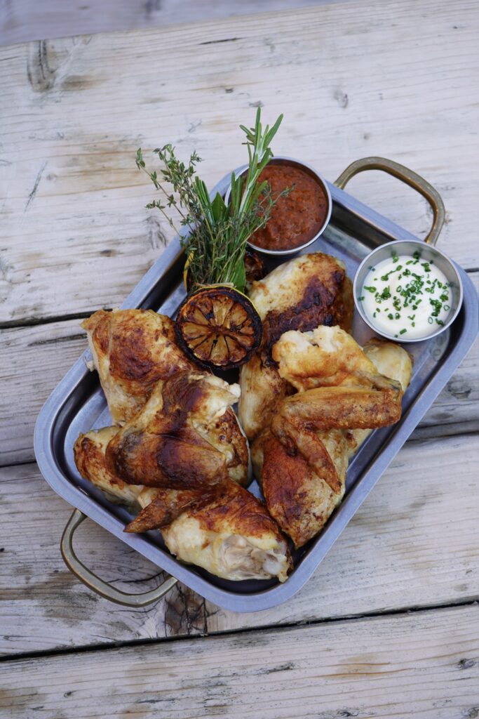 Rotisserie chicken pieces in a steel tray with charred lemon, fresh herbs and two dips, on a wooden table